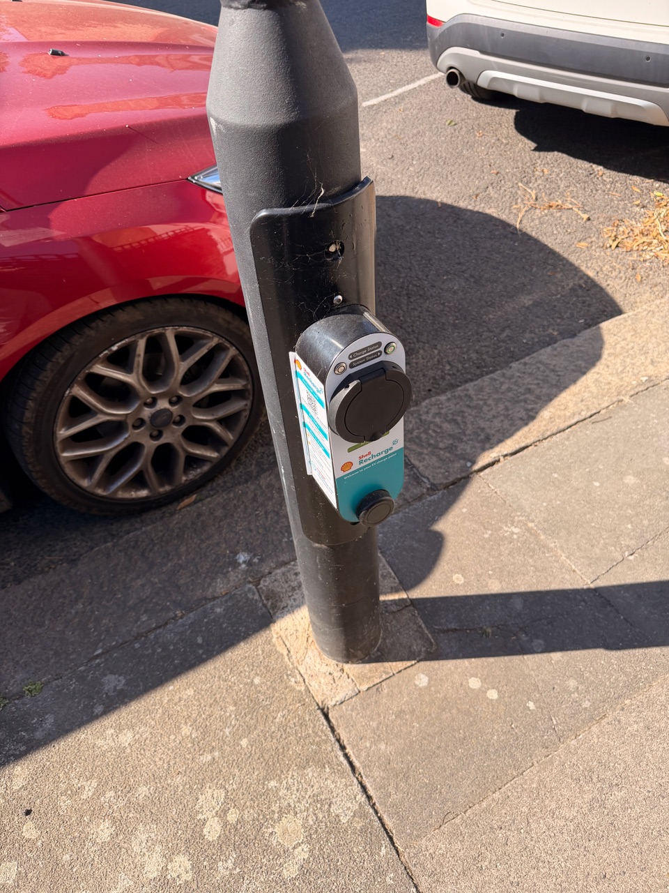 Kompakte Ladestation für Elektroautos, die an einem schwarzen Straßenlaternenmast befestigt ist. Daneben parkt ein rotes Auto; im Hintergrund ist ein weiteres Fahrzeug zu sehen.