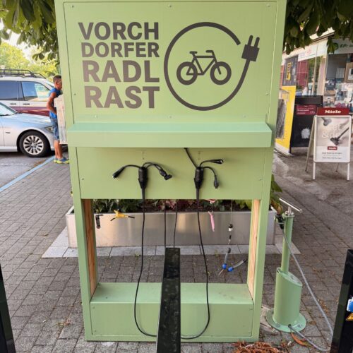 Grüne öffentliche E-Bike-Ladestation mit der Aufschrift „Vorchdorfer Radl Rast“. Mehrere Ladekabel mit unterschiedlichen Steckern hängen an der Station, daneben befindet sich eine Fahrrad-Servicestelle. Die Anlage steht im Ortszentrum unter einem Baum.