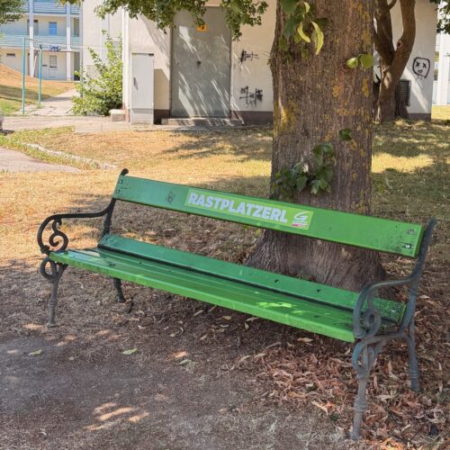 Grün gestrichene Parkbank mit der Aufschrift „RASTPLATZERL“ unter einem großen schattenspendenden Baum. Im Hintergrund stehen Wohngebäude; der Rasen rund um die Bank ist vollkommen ausgetrocknet.